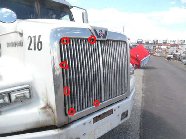 A white semi-truck with visible damage and red markings indicating points of interest, parked in a lot with other vehicles and parts in the background.