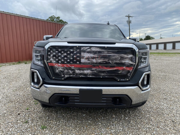 A black pickup truck with a Bug Screen: Smokey Thin Red Line grille insert parked in front of a red building under a cloudy sky.