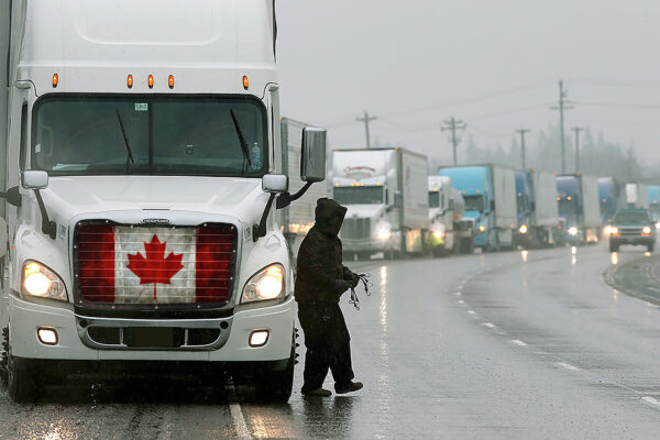 A person stands in the rain beside a truck with a Canadian Flag - Semi Truck Mesh Bug Screen on the front, with a lineup of trucks in the background.