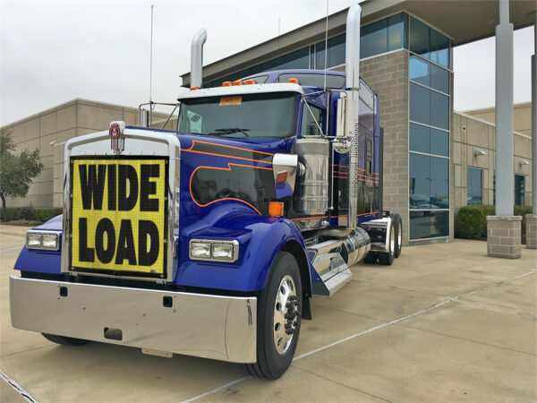 Blue semi-truck equipped with a Bug Screen: Wide Load sign parked in front of a building.